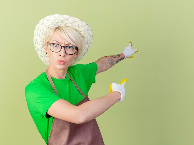 Jeune jardinier femme aux cheveux courts en tablier et chapeau portant des gants en caoutchouc pointant vers quelque chose en arrière avec l'index à la confusion debout sur fond clair