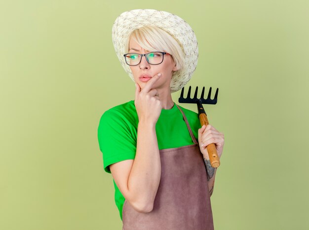 Jeune jardinier femme aux cheveux courts en tablier et hat holding mini rake à côté perplexe debout sur fond clair