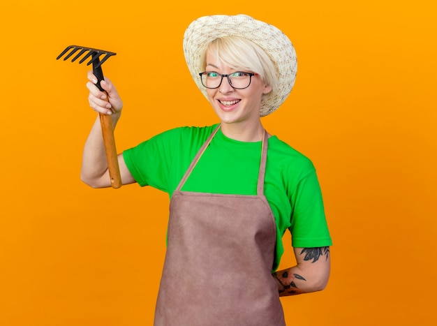 Jeune jardinier femme aux cheveux courts en tablier et hat holding mini rake looking at camera smiling joyeusement debout sur fond orange