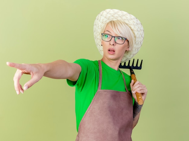 Jeune jardinier femme aux cheveux courts en tablier et hat holding mini rake pointant vers quelque chose avec l'index à la surprise debout sur fond clair