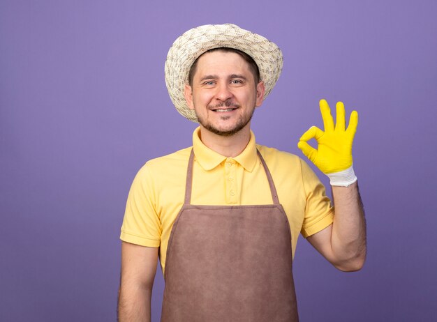 Jeune jardinier homme portant une combinaison et un chapeau dans des gants de travail à l'avant souriant joyeusement montrant signe ok debout sur le mur violet