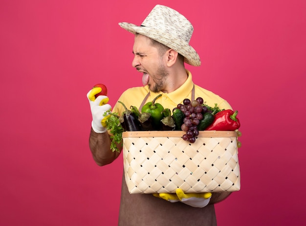 Jeune jardinier homme portant une combinaison et un chapeau dans des gants de travail tenant une caisse pleine de légumes à la tomate fraîche à la main avec une expression dégoûtée