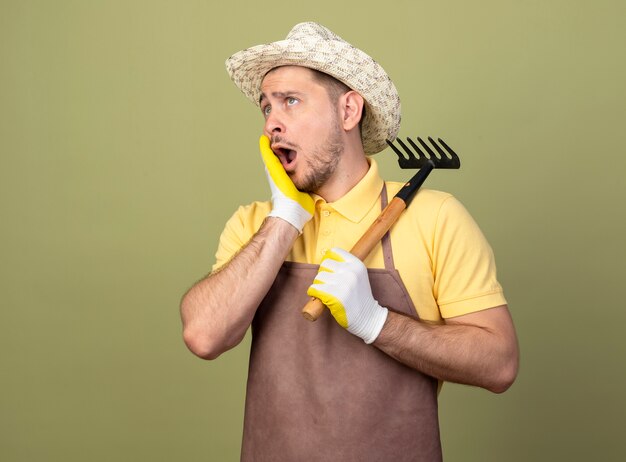 Jeune jardinier homme portant une combinaison et un chapeau dans des gants de travail tenant un mini râteau à côté étonné et surpris debout sur un mur léger