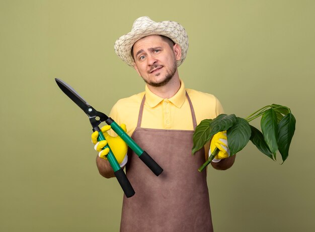 Jeune jardinier homme portant une combinaison et un chapeau dans des gants de travail tenant une plante et un coupe-haie souriant avec un visage heureux