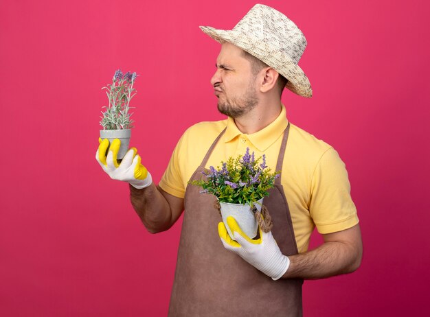 Jeune jardinier homme portant une combinaison et un chapeau dans des gants de travail tenant des plantes en pot en les regardant être confus et mécontent