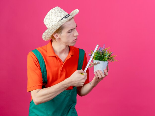 Jeune jardinier homme portant une combinaison et un chapeau tenant une plante en pot le mesurant
