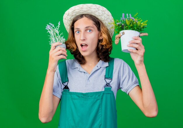 Jeune jardinier homme portant combinaison et chapeau tenant des plantes en pot surpris