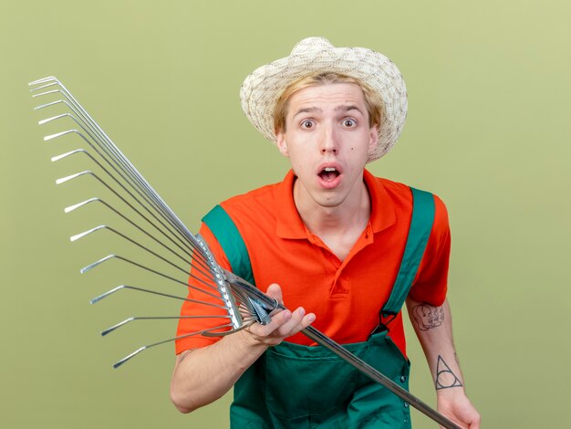 Jeune jardinier homme portant combinaison et chapeau tenant râteau regardant la caméra étonné et surpris debout sur fond clair