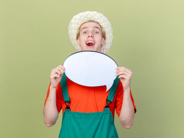 Jeune jardinier homme portant une combinaison et un chapeau tenant un signe de bulle de discours vierge furtivement sur souriant joyeusement debout sur fond clair