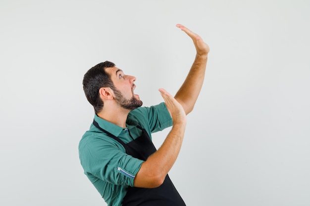 Jeune jardinier levant les mains de manière protectrice en t-shirt, tablier et ayant l'air effrayé. vue de face.