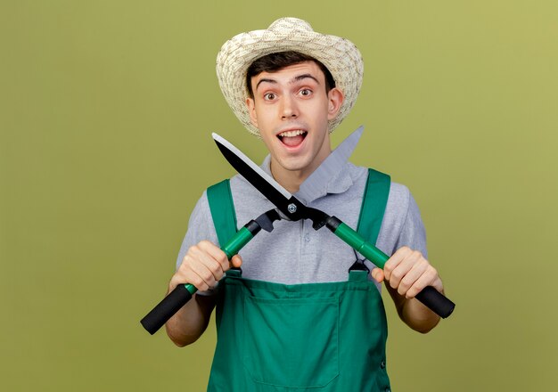 Le jeune jardinier masculin enthousiaste portant le chapeau de jardinage tient des tondeuses