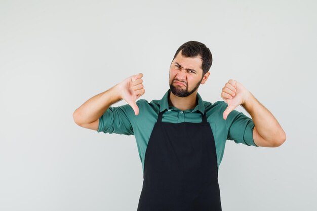 Jeune jardinier montrant le pouce vers le bas en t-shirt, tablier et l'air mécontent. vue de face.