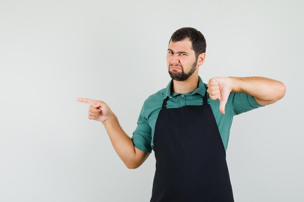 Jeune jardinier pointant de côté tout en montrant le pouce vers le bas en t-shirt, tablier et l'air insatisfait, vue de face.