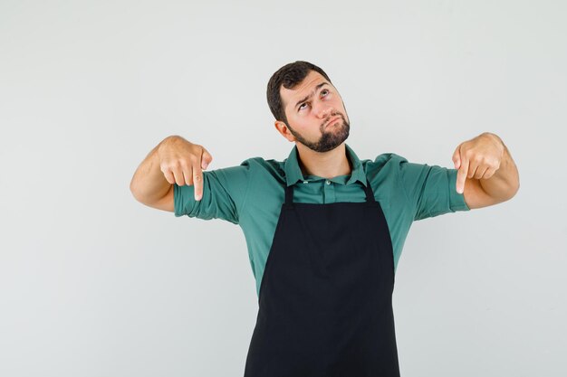 Jeune jardinier pointant vers le bas en t-shirt, tablier et ayant l'air confus. vue de face.