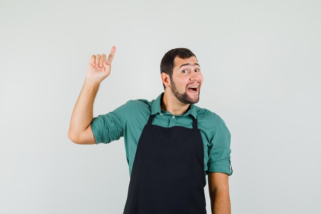 Jeune jardinier pointant vers le haut en t-shirt, tablier et semblant joyeux. vue de face.