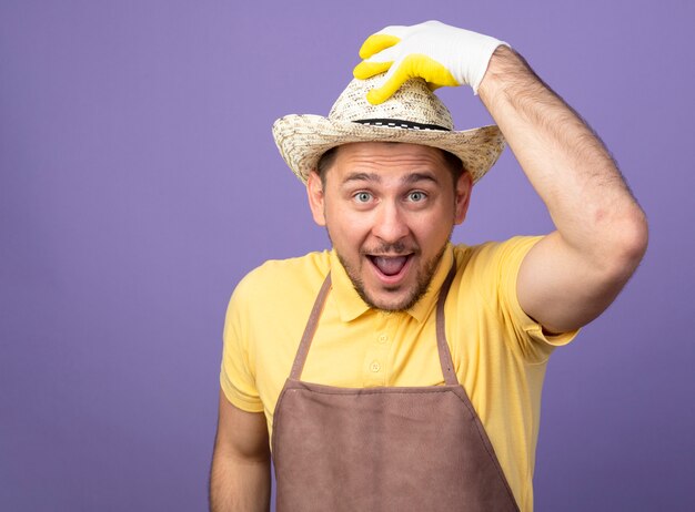 Jeune jardinier portant combinaison et chapeau dans des gants de travail à l'avant souriant joyeux toucher son chapeau debout sur le mur violet