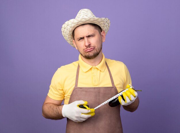 Jeune jardinier portant combinaison et chapeau dans des gants de travail tenant l'aubergine le mesurant avec un ruban à mesurer à la confusion debout sur le mur violet