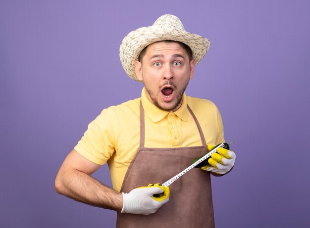 Jeune jardinier portant combinaison et chapeau dans des gants de travail tenant l'aubergine en le mesurant avec un ruban à mesurer à la surprise debout sur le mur violet