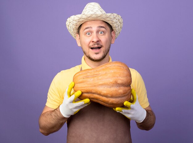 Jeune jardinier portant combinaison et chapeau dans des gants de travail tenant la citrouille à l'avant souriant avec un visage heureux debout sur un mur violet