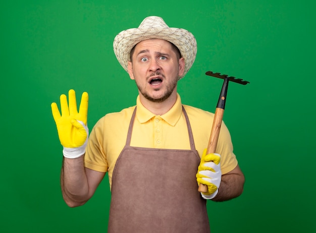 Jeune jardinier portant combinaison et chapeau dans des gants de travail tenant un mini râteau à l'avant montrant confus numéro quatre debout sur un mur vert