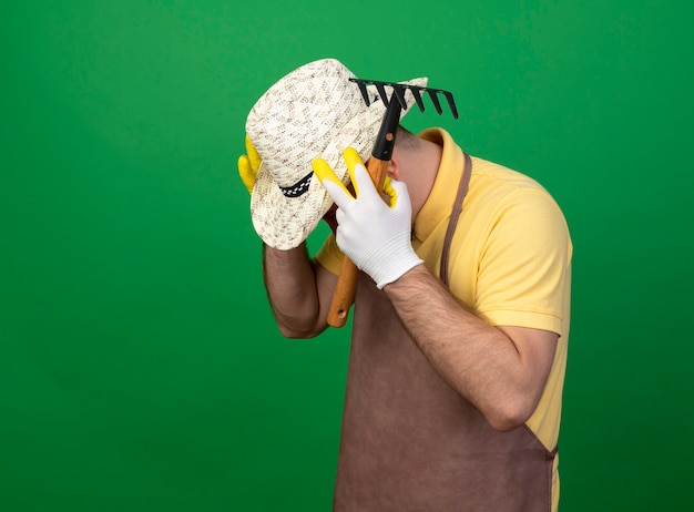 Jeune jardinier portant combinaison et chapeau dans des gants de travail tenant un mini râteau à la main dans la main frustré sur sa tête debout sur un mur vert