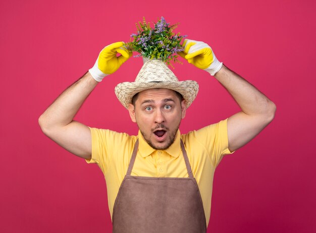 Jeune jardinier portant une combinaison et un chapeau dans des gants de travail tenant une plante en pot au-dessus de sa tête à la surprise et surpris debout sur le mur rose