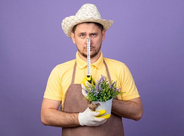 Photo gratuite jeune jardinier portant une combinaison et un chapeau dans des gants de travail tenant une plante en pot et un ruban à mesurer à l'avant avec un visage sérieux debout sur un mur violet