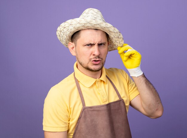 Jeune jardinier portant combinaison et chapeau en gants de travail à l'avant avec une expression confiante sérieuse debout sur un mur violet