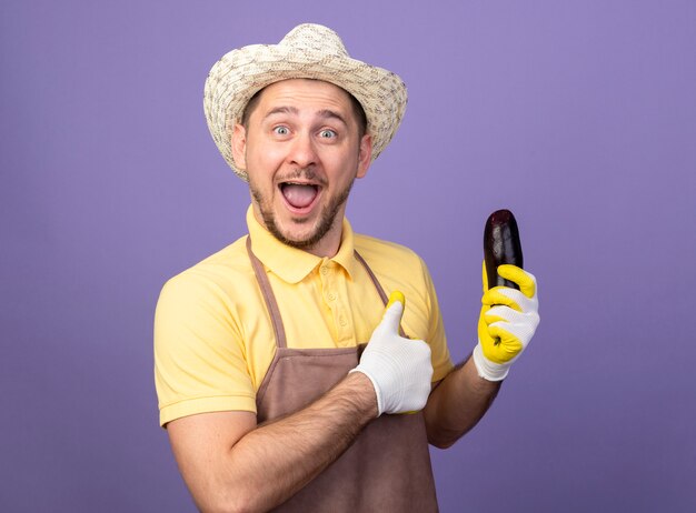 Jeune jardinier portant combinaison et chapeau en gants de travail tenant l'aubergine à l'avant montrant les pouces vers le haut debout sur le mur violet