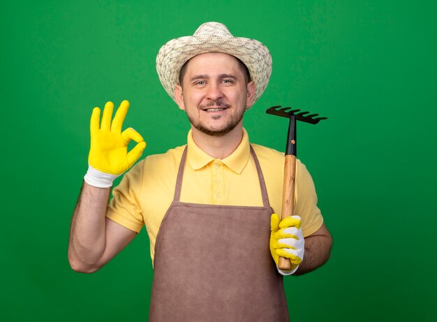 Jeune jardinier portant combinaison et chapeau en gants de travail tenant mini râteau à l'avant souriant montrant signe ok debout sur mur vert
