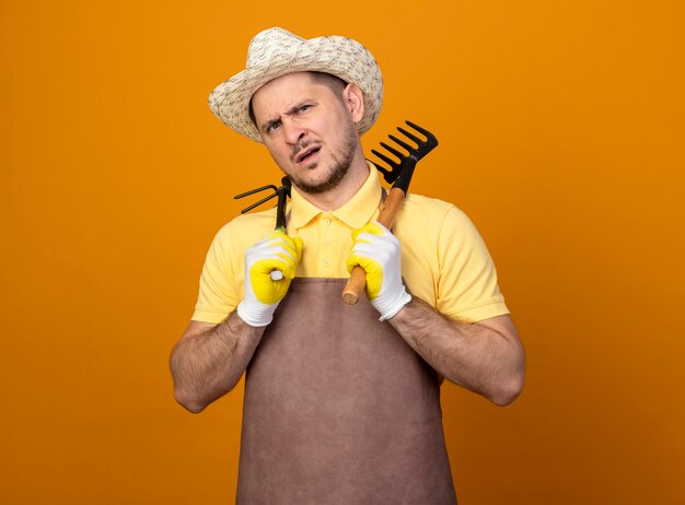 Jeune jardinier portant combinaison et chapeau tenant Mattock et mini râteau à l'avant avec une expression sceptique debout sur un mur orange