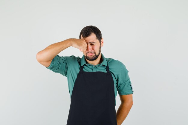 Jeune jardinier se frottant les yeux en pleurant en t-shirt, en tablier et en ayant l'air triste. vue de face.