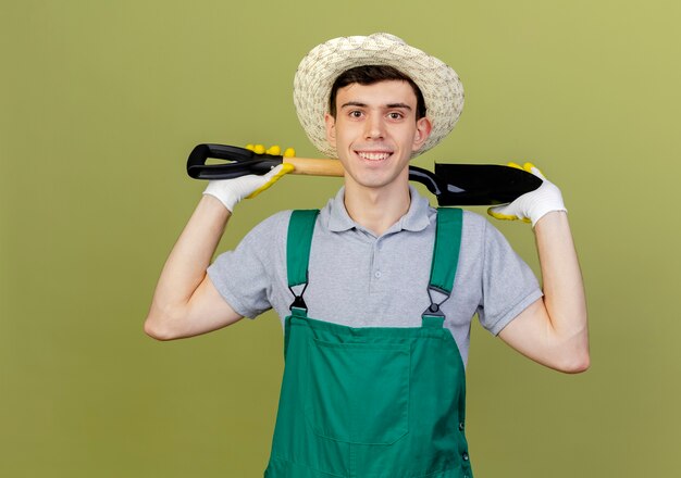 Un jeune jardinier souriant portant un chapeau et des gants de jardinage tient une pelle derrière le cou