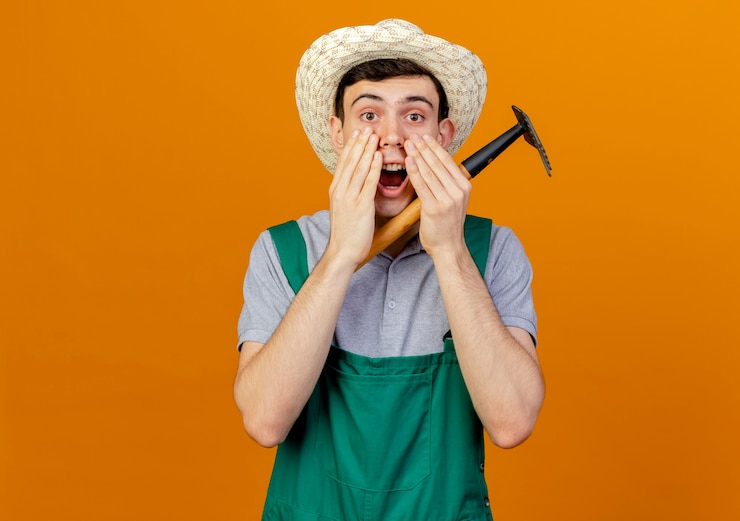 Photo gratuit un jeune jardinier surpris portant un chapeau de jardinage met les mains sur la bouche tenant un râteau