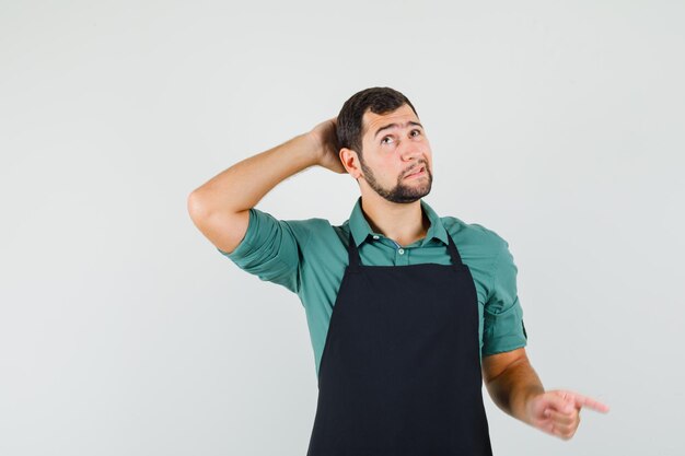 Jeune jardinier en t-shirt, tablier tenant la main sur sa tête tout en pointant de côté et l'air perplexe, vue de face.