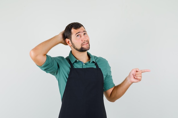 Jeune jardinier tenant la main sur sa tête tout en pointant de côté en t-shirt, tablier et à la vue de face perplexe