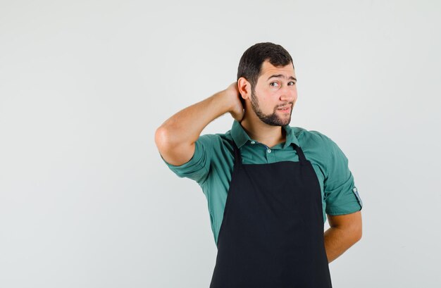 Jeune jardinier tenant la main sur son cou en t-shirt, tablier et semblant attrayant, vue de face.