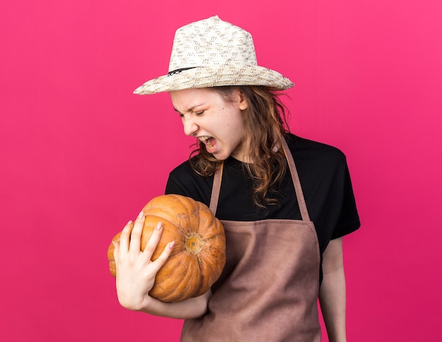 Jeune jardinière en colère portant un chapeau de jardinage tenant et regardant la citrouille