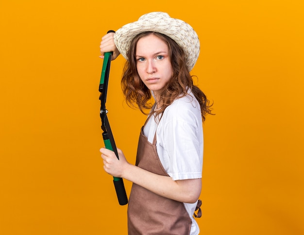 Jeune jardinière confiante portant un chapeau de jardinage tenant un sécateur isolé sur un mur orange