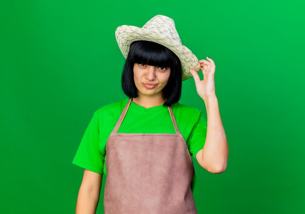 Jeune jardinière confiante en uniforme portant et tenant un chapeau de jardinage isolé