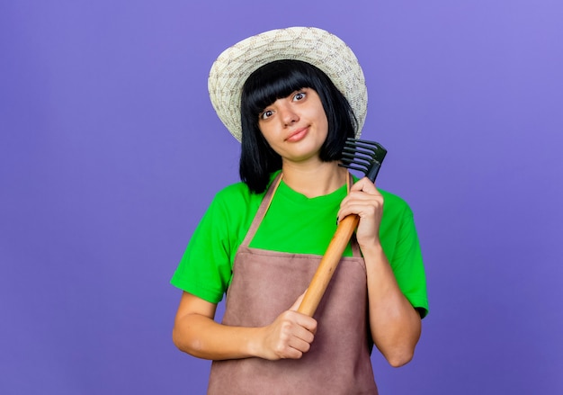 Jeune jardinière enthousiaste en uniforme portant un chapeau de jardinage détient un râteau