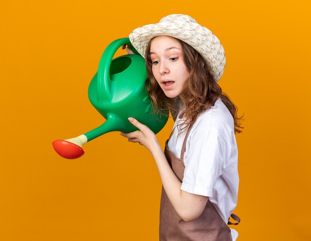 Photo gratuite jeune jardinière impressionnée portant un chapeau de jardinage arrosant avec un arrosoir isolé sur un mur orange