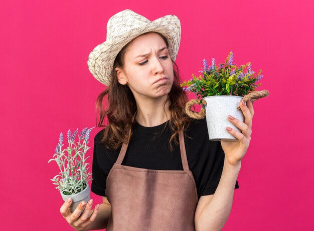 Jeune jardinière mécontente portant un chapeau de jardinage tenant et regardant des fleurs dans des pots de fleurs isolés sur un mur rose