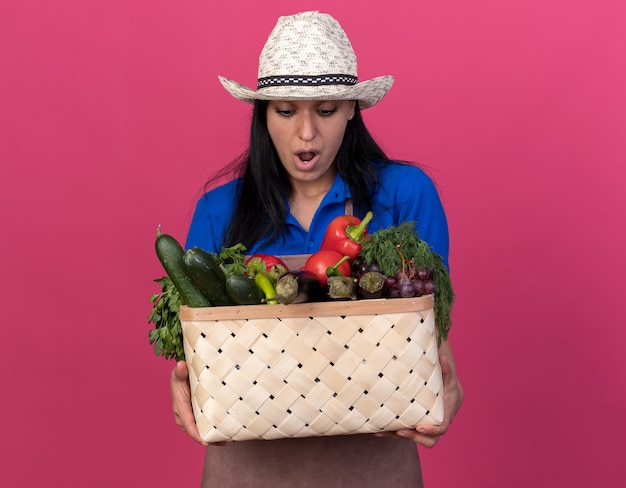 Jeune jardinière surprise en uniforme et chapeau tenant et regardant un panier de légumes isolé sur un mur rose avec espace pour copie