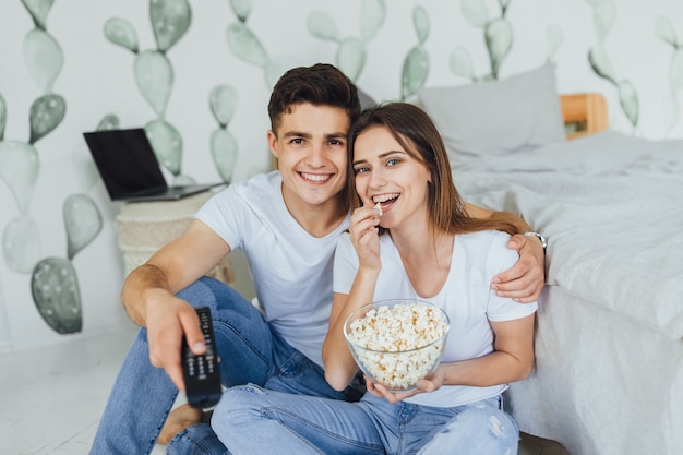 Jeune joli couple en vêtements décontractés regardant la télévision à la maison au chevet et mangeant du pop-corn
