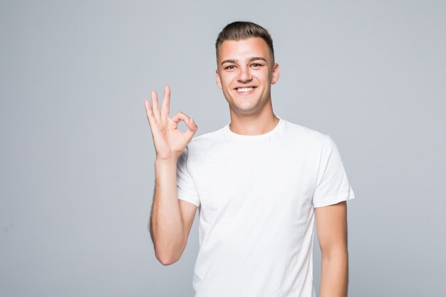 Jeune joli homme dans un T-shirt blanc isolé sur blanc montre signe ok