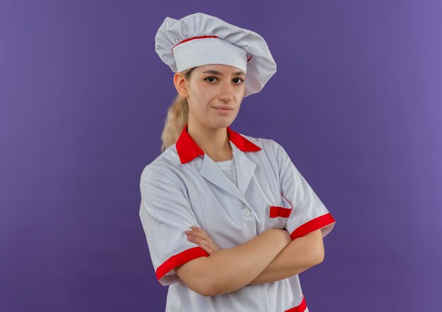 Jeune jolie cuisinière confiante en uniforme de chef debout avec une posture fermée, isolée sur un mur violet avec espace pour copie