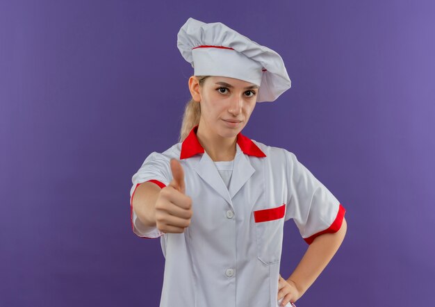 Jeune jolie cuisinière confiante en uniforme de chef mettant la main sur la taille et montrant le pouce vers le haut isolé sur un mur violet avec espace de copie