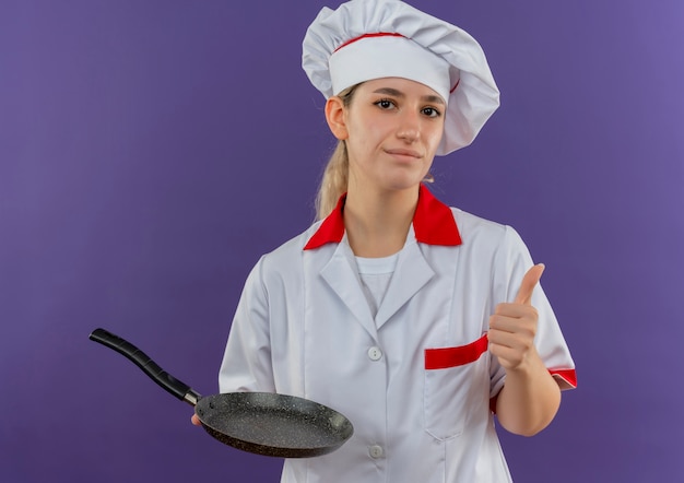 Jeune jolie cuisinière confiante en uniforme de chef tenant une poêle à frire et montrant le pouce vers le haut isolé sur un mur violet