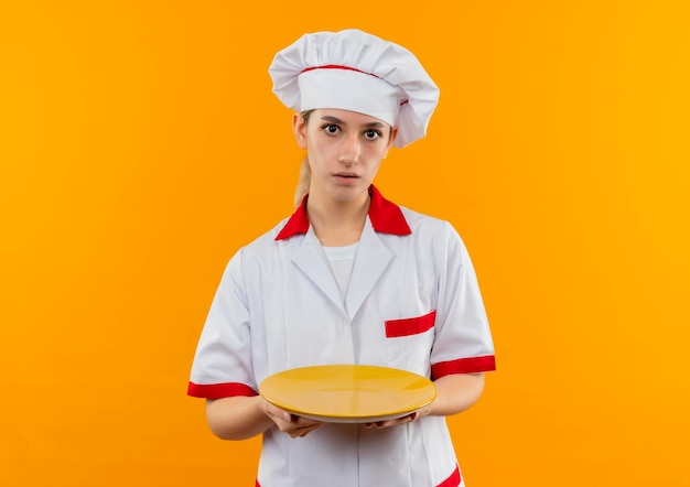 Jeune jolie cuisinière impressionnée en uniforme de chef tenant une assiette vide isolée sur un mur orange